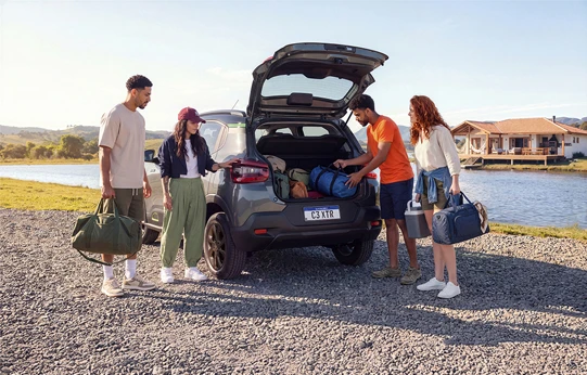 Uma mulher de cabelo preso, camiseta verde e macacão jeans coloca seus itens de viagem no porta-malas do Citroën C3 XTR, ao lado de uma piscina, em um dia ensolarado.