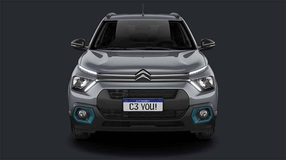 Design e Atitude Parte frontal do Citroën C3 prata com logotipo Citroën na grade, luzes DRL, faróis e placa "C3 YOU!" visíveis.