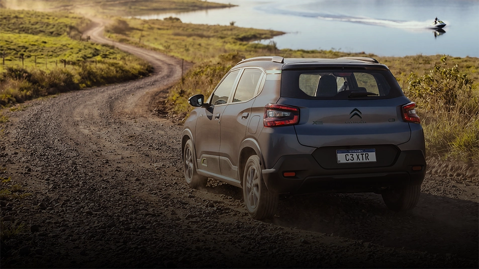 Suspensão com efeito tapete voador Citroën C3 XTR cinza com teto preto, parte traseira e lateral visíveis, circulando em estrada de terra com vegetação ao redor.
