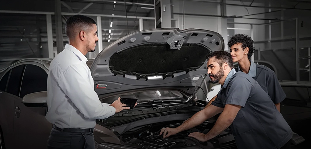 Com o capô do veículo aberto, duas pessoas uniformizadas inspecionam o motor, ao lado de um homem de camisa branca. 
