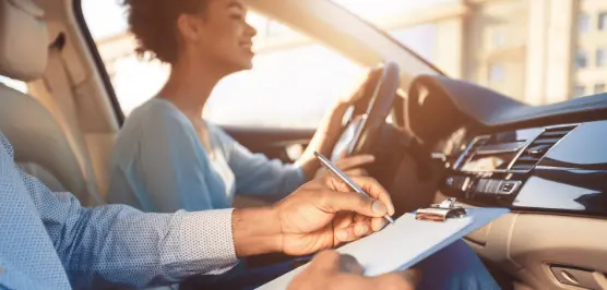 Duas pessoas com tom de pele morena dentro de um carro. Na condução, uma mulher com cabelo curto encaracolado dirige e no passageiro um homem faz anotações em uma prancheta&nbsp;&nbsp;